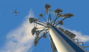 High quality 3D render of a UAV drone inspecting a cellular phone tower. Fictitious UAV and wireless tower antenna array; slightly overcast blue sky and motion blur for dramatic effect.